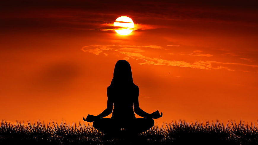 Woman meditating in lotus pose silhouette against vibrant orange sunset sky