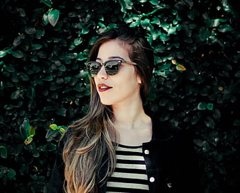 Stylish young woman wearing sunglasses and striped top against green ivy wall