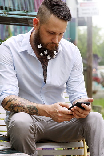 Tattooed Man with Flowers in Beard Using Smartphone