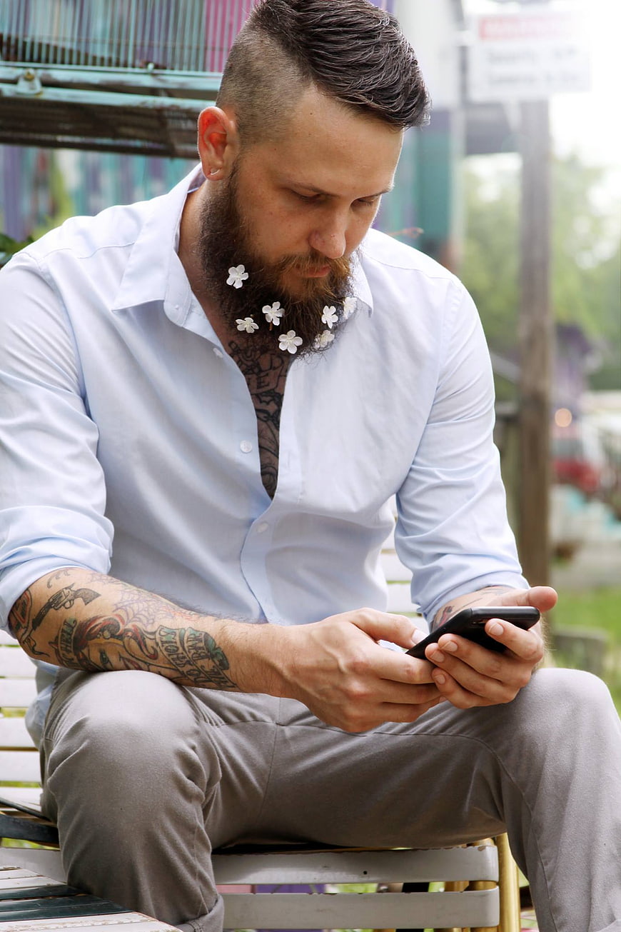 Tattooed Man with Flowers in Beard Using Smartphone