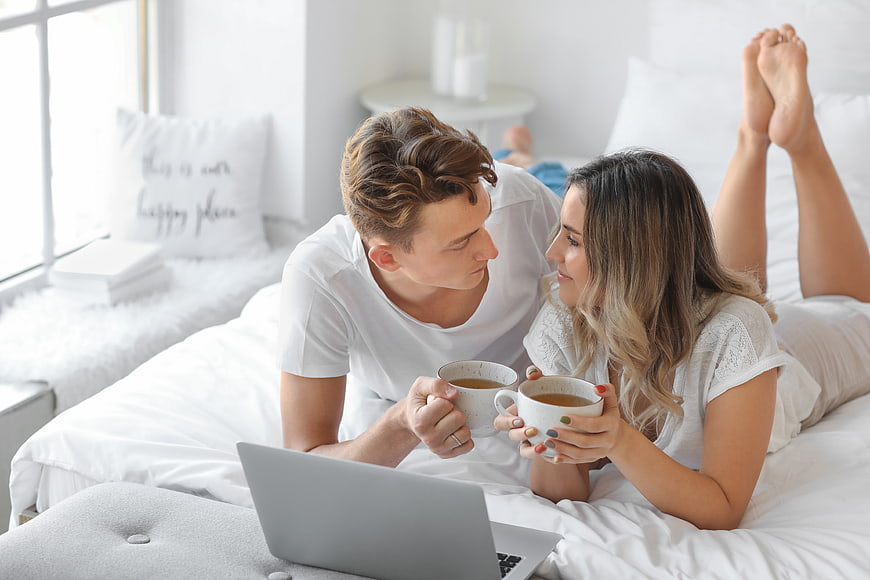 Happy couple drinking coffee in bed with laptop