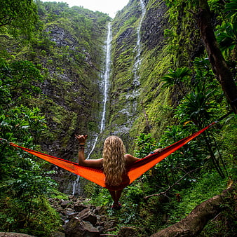 Woman relaxing in orange hammock facing lush waterfall
