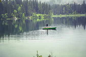 Lone fisherman in green boat on calm lake with forest reflection