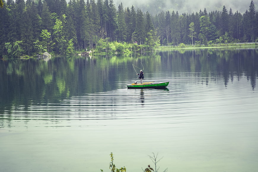 Lone fisherman in green boat on calm lake with forest reflection
