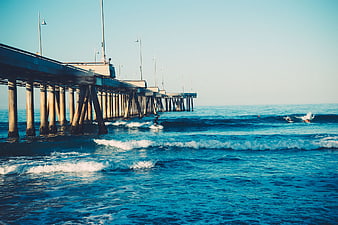 Surfer riding waves near a long concrete pier on a sunny day