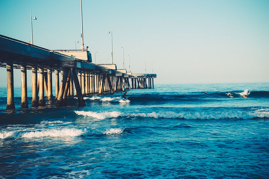 Surfer riding waves near a long concrete pier on a sunny day