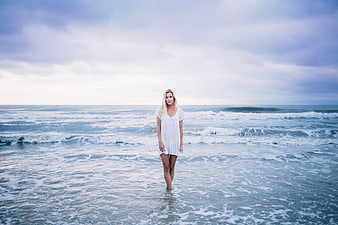 Blonde woman in white dress standing in ocean waves on a cloudy beach