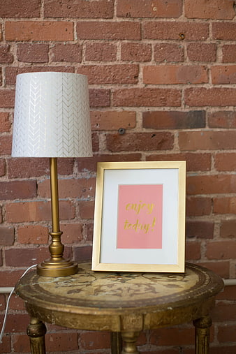 Vintage gold lamp and framed 'enjoy today' print on table against brick wall