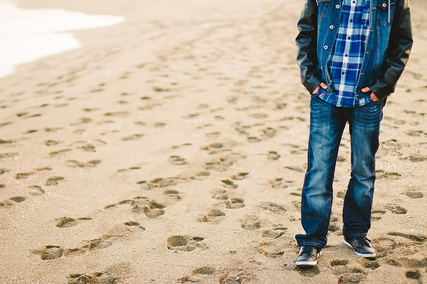 Man in Denim Standing on Sandy Beach with Footprints