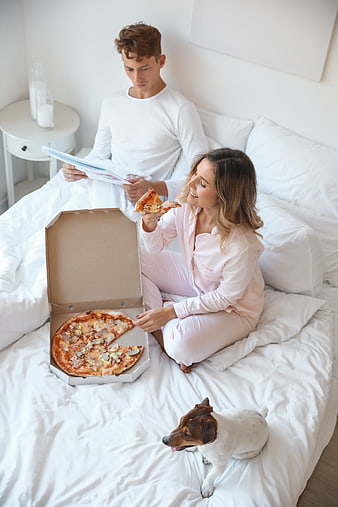 Young couple eating pizza in bed with dog
