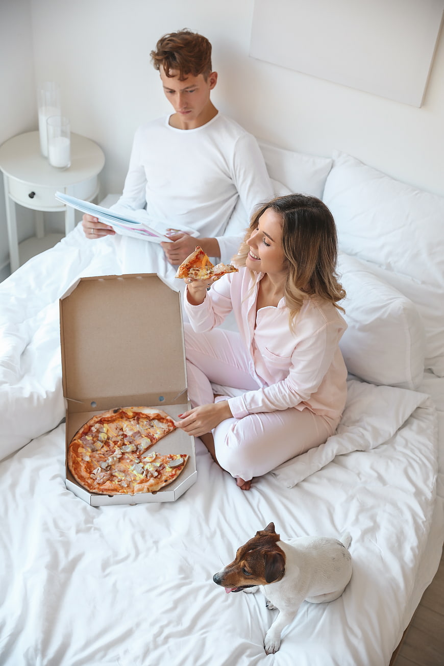 Young couple eating pizza in bed with dog