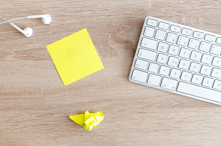 Flat lay of white keyboard, yellow sticky note, and earphones on wooden desk