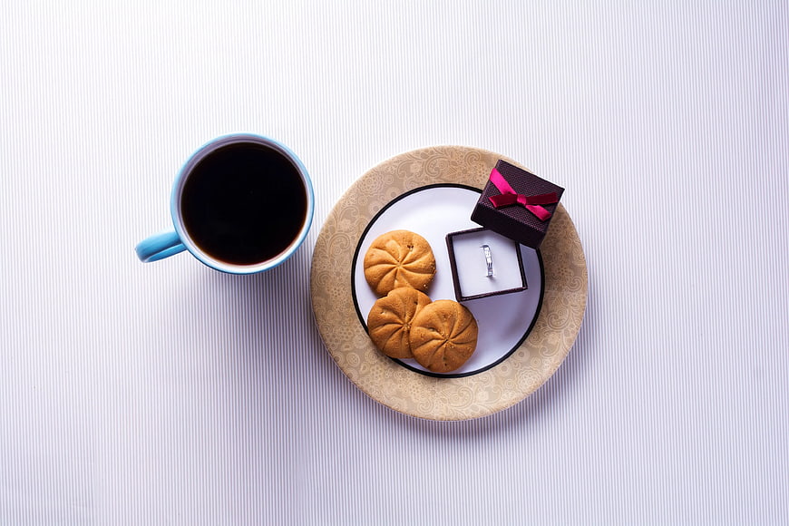 Top view of coffee, cookies, and engagement ring on white table