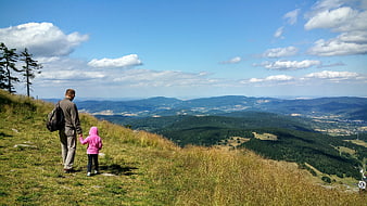 Father and daughter holding hands on a grassy mountain ridge overlooking a vast valley