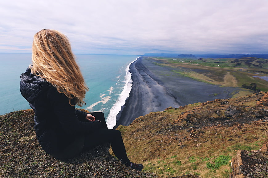 Woman sitting on cliff overlooking black sand beach and ocean
