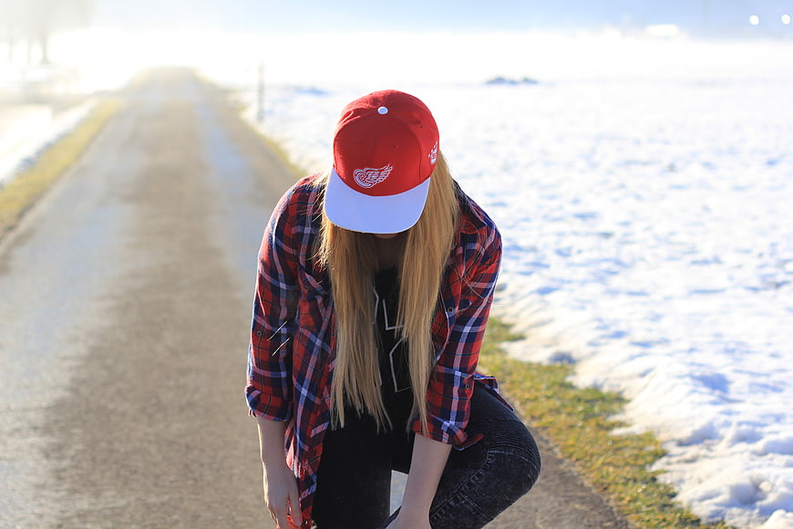 Woman in Red Cap and Plaid Shirt Crouching on Snowy Road