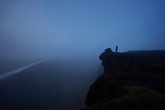 Silhouette of Man Standing on Cliff Edge in Foggy Blue Landscape