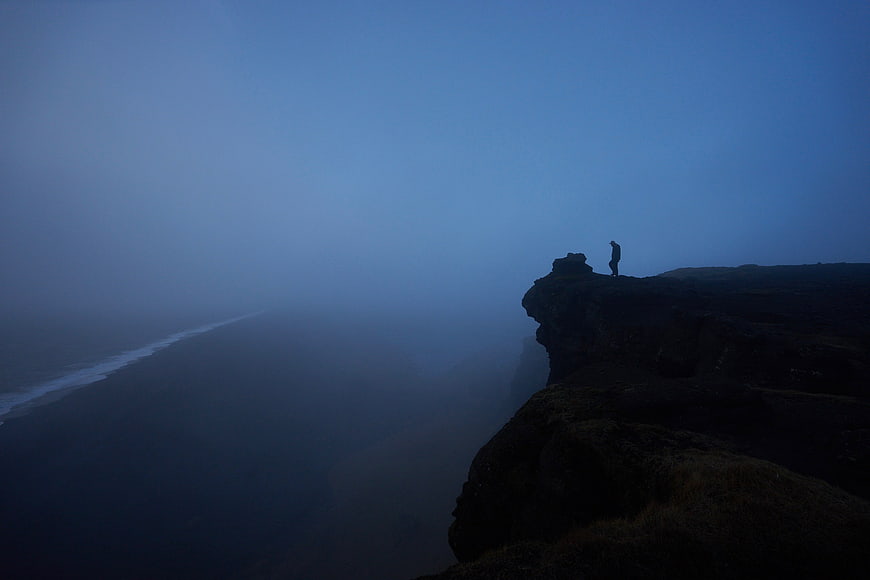Silhouette of Man Standing on Cliff Edge in Foggy Blue Landscape