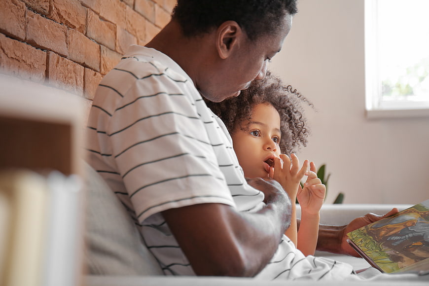 Father reading storybook to young daughter on couch