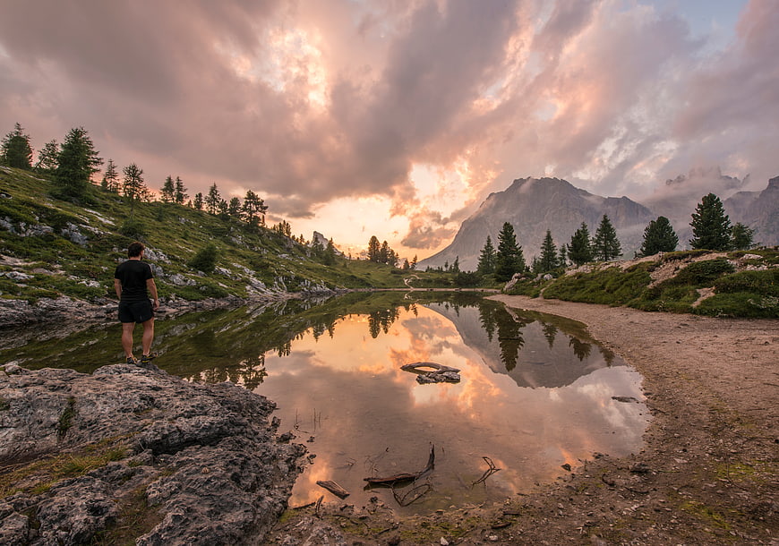 Man standing by alpine lake at sunset with mountain reflection
