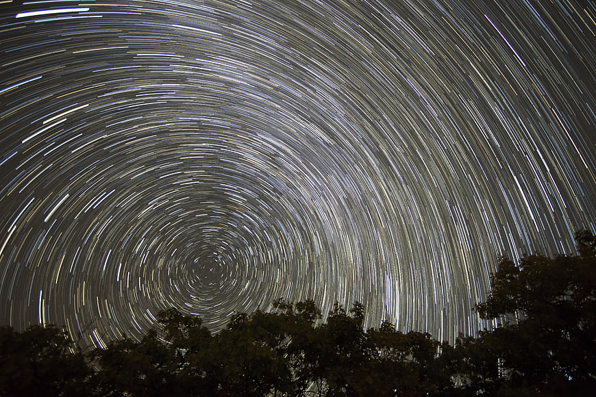 Star trails circling the night sky above a dark forest silhouette