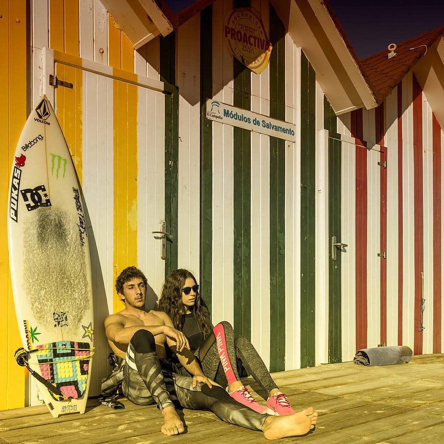 Couple relaxing by surfboard in front of colorful beach huts