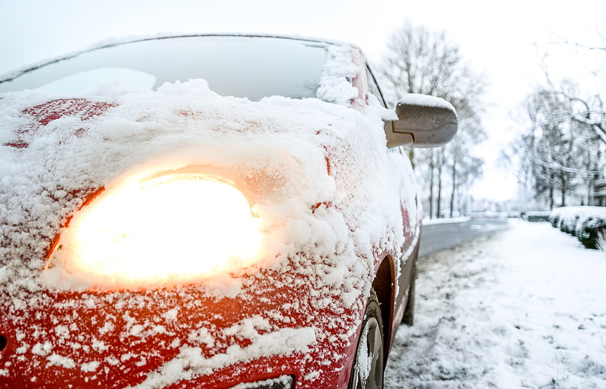 Red car with headlights on covered in snow