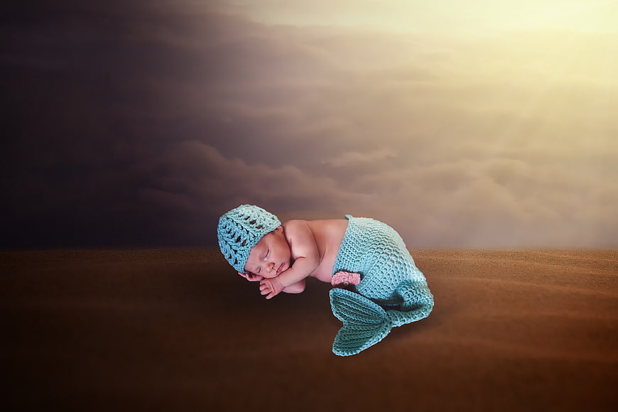 Newborn baby sleeping in mermaid costume against dramatic desert sky