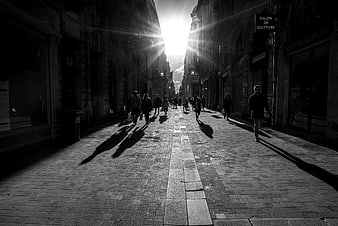 Black and white street photography of pedestrians walking down a cobblestone alley with dramatic sunlight and long shadows.