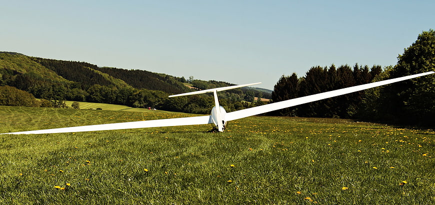 White glider aircraft parked in a green meadow with forested hills