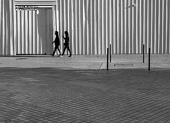 Two People Walking Past Corrugated Metal Wall in Black and White