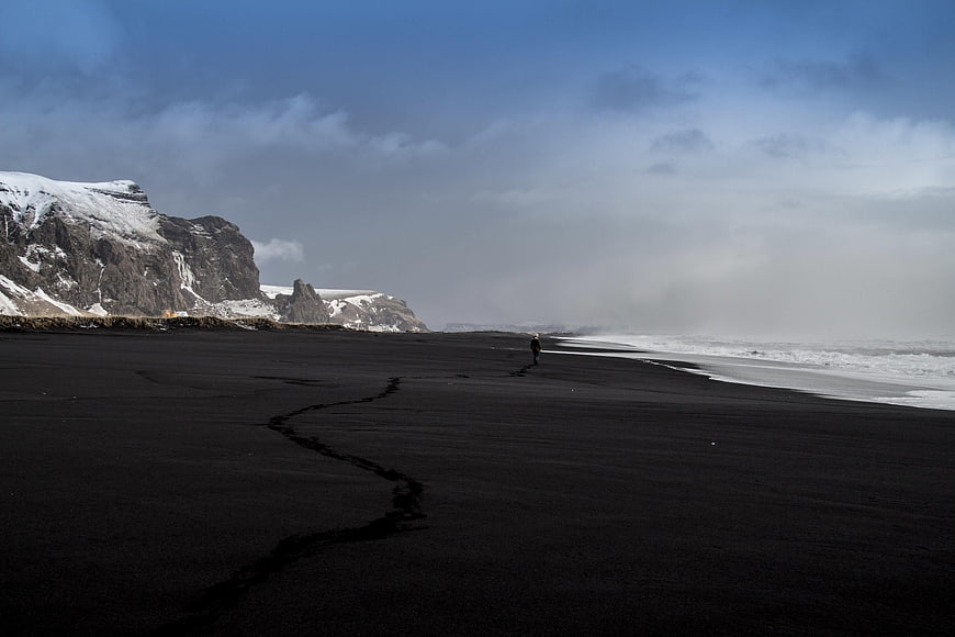 Lone Figure Walking on Black Sand Beach with Snowy Cliffs