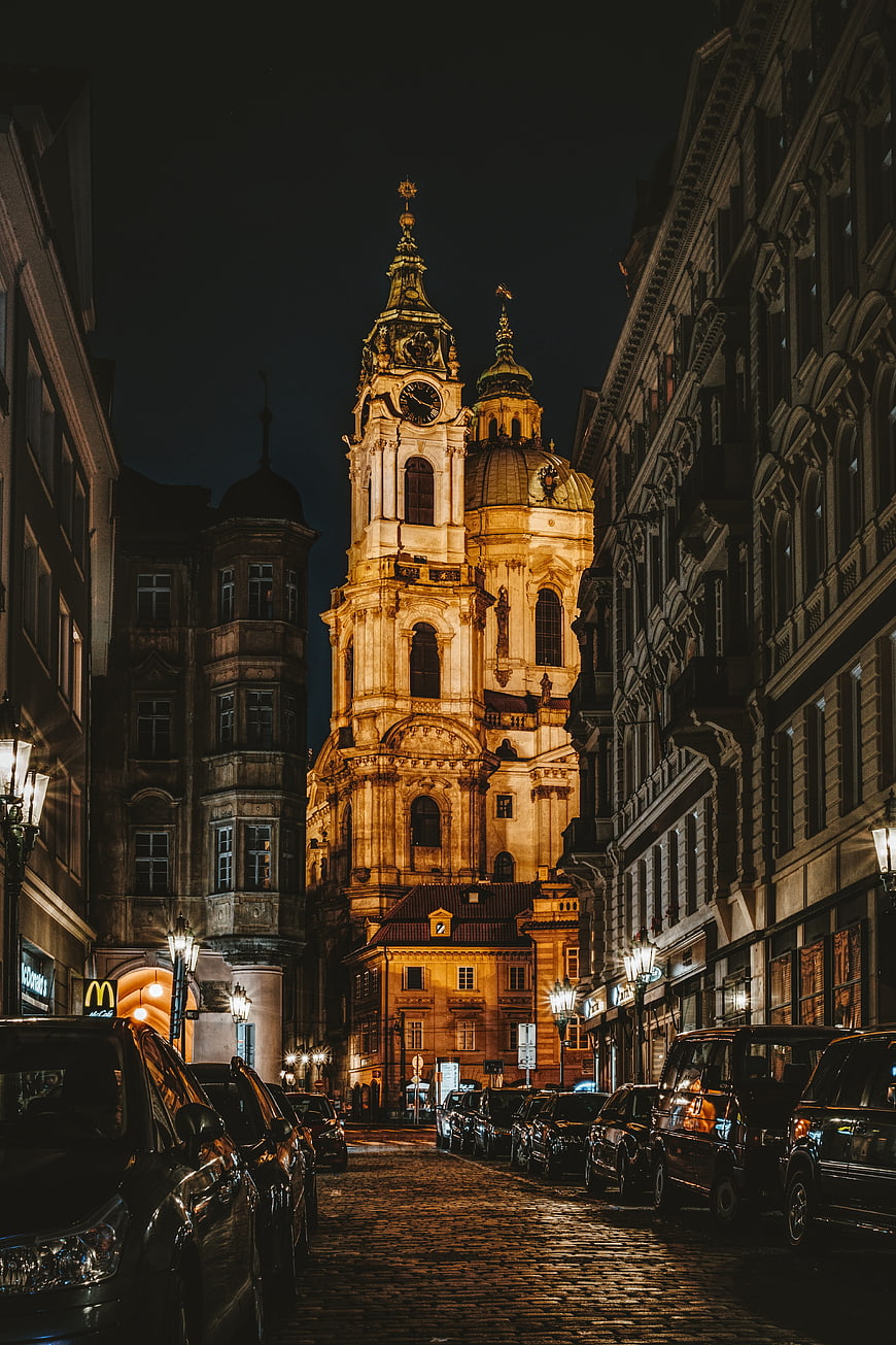Illuminated Baroque Church Tower on Cobblestone Street in Prague at Night