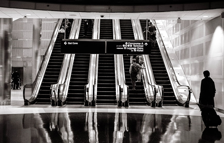 Black and white airport escalators with travelers and directional signs