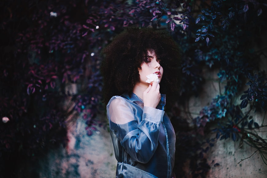 Woman with Curly Hair Holding White Flower Against Purple Foliage