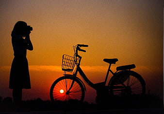 Silhouette of woman photographer and bicycle against orange sunset sky