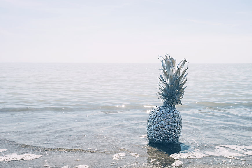 Surreal silver pineapple standing in ocean waves on beach