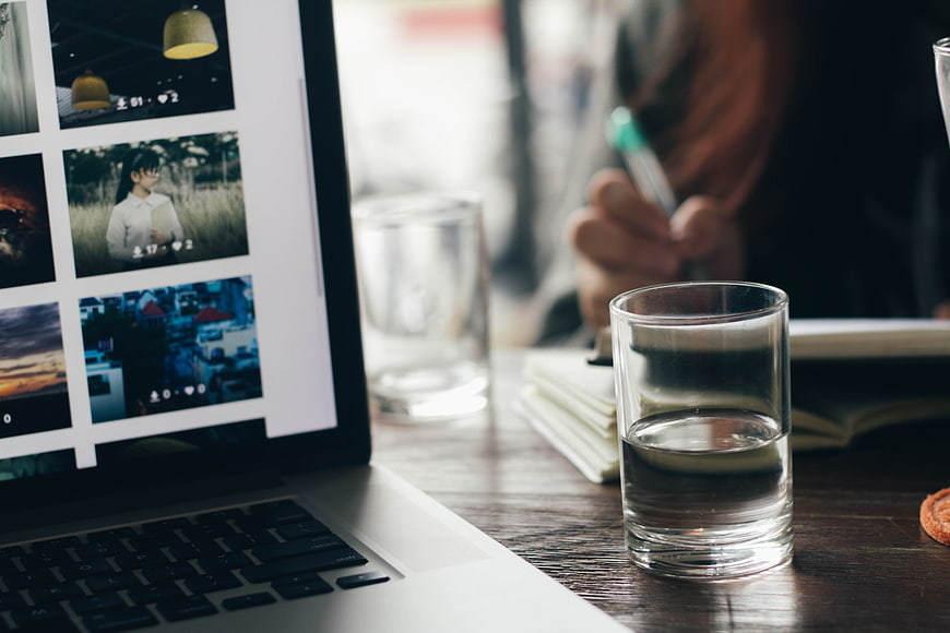 Laptop displaying photo grid on wooden desk with water glass