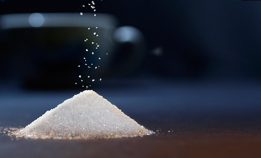 Pile of white sugar with falling granules and blurred coffee cup