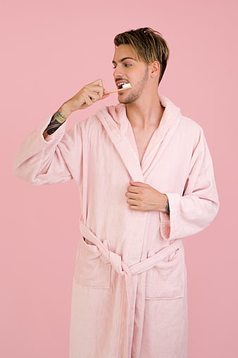 Handsome man in pink bathrobe brushing teeth on pink background