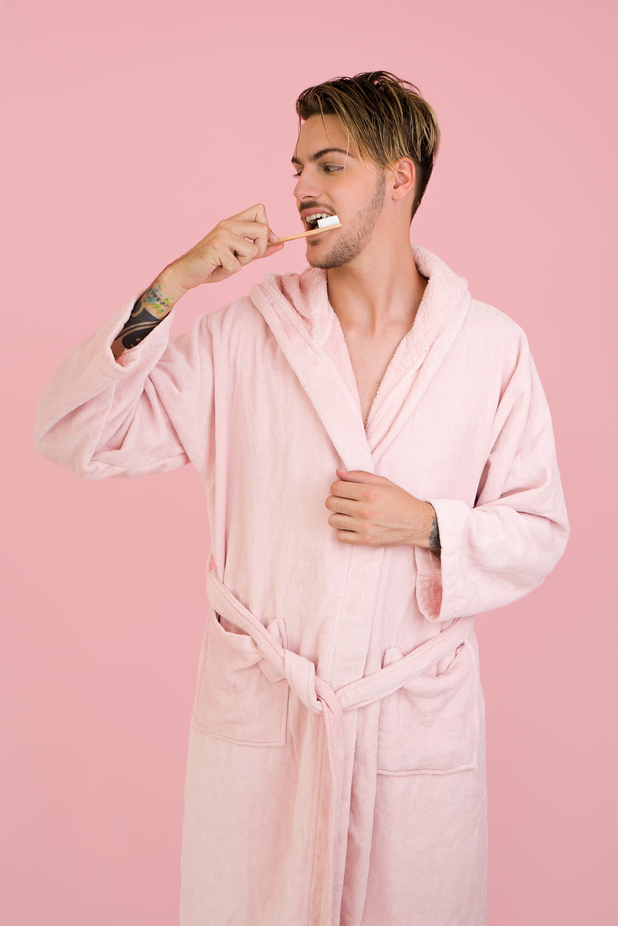 Handsome man in pink bathrobe brushing teeth on pink background