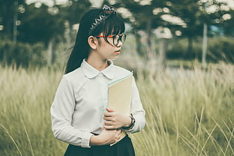Asian student girl with glasses holding folder in grassy field