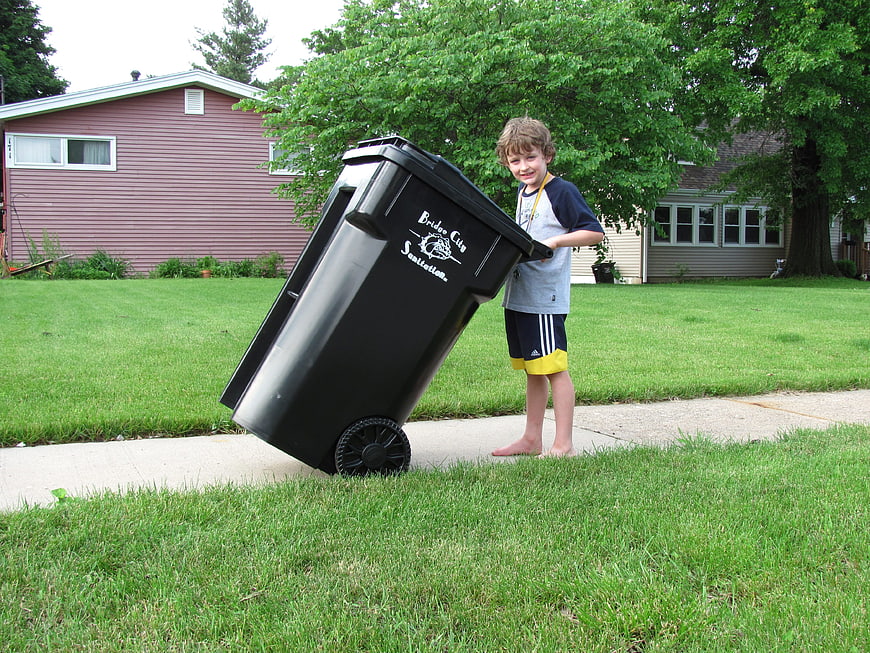 Young boy pushing large black garbage bin on suburban sidewalk