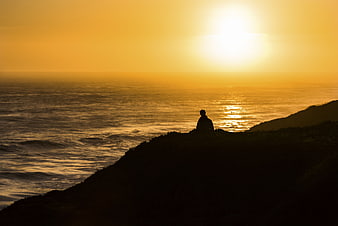 Silhouette of Man Sitting on Cliff Watching Golden Sunset Over Ocean