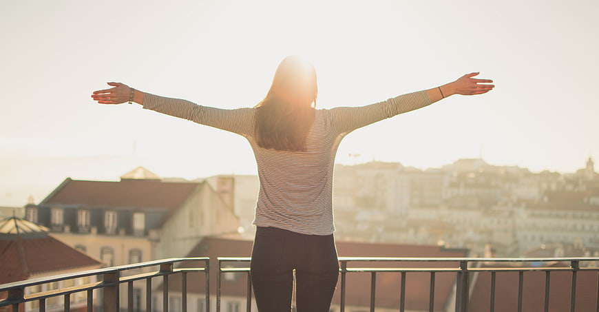 Woman with arms raised on balcony overlooking city at sunset