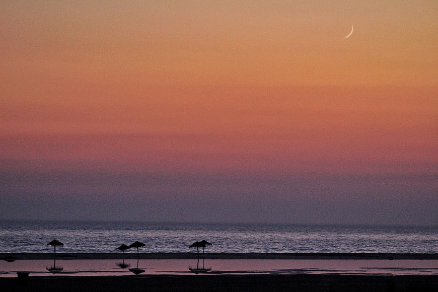 Silhouetted beach umbrellas at sunset with crescent moon