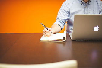 Man Writing in Notebook with Laptop on Desk