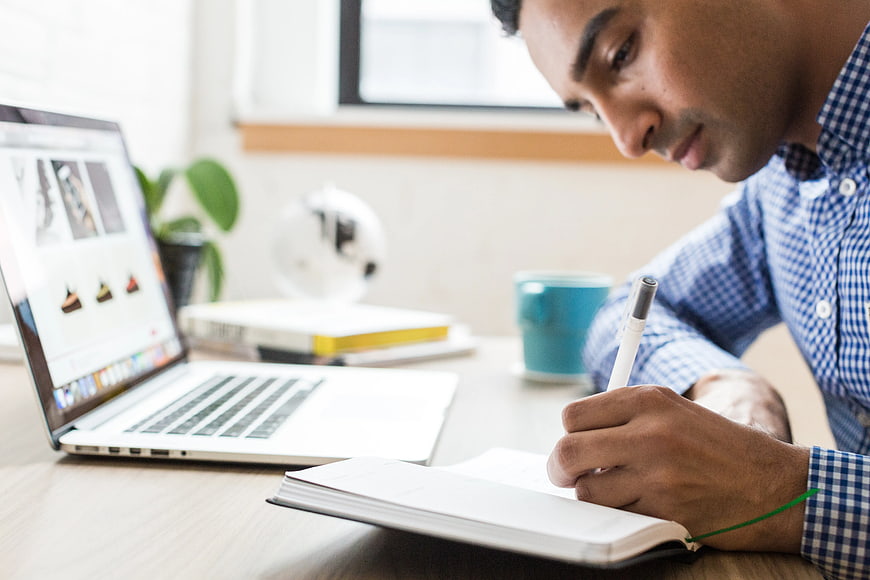 Man Writing in Notebook at Desk with Laptop