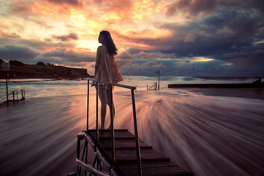 Woman standing on pier looking at dramatic sunset over ocean