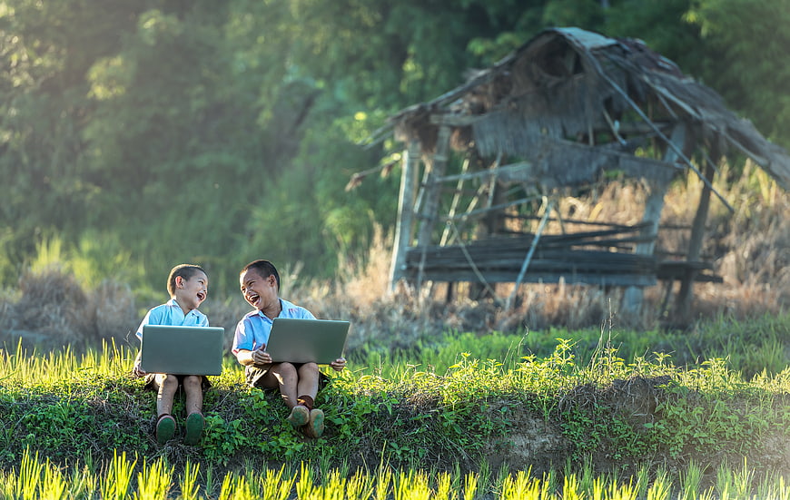 Two laughing boys using laptops in a rural rice field